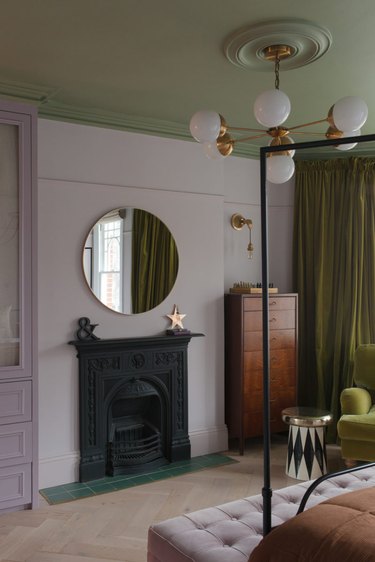 Bedroom with lavender walls, green ceiling and curtains, brown bedding and a black painted fireplace.