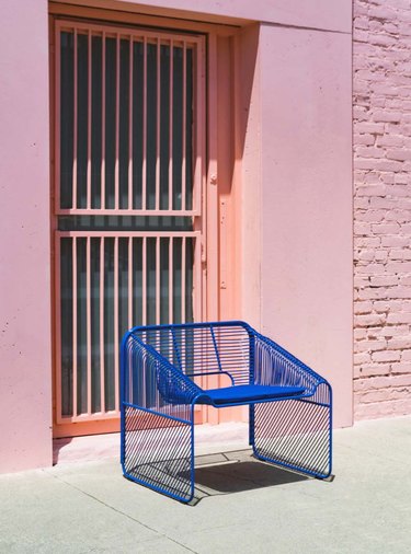 Blue wire chair is angled outdoors on sidewalk in front of melon door gate and pink entryway of pink brick building
