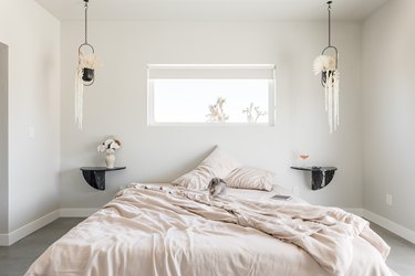 Minimalist bedroom with beige bedding, twin black shelf nightstands, double hanging planters, and rectangular window