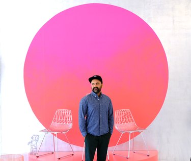 Man with dark beard wearing black baseball cap, blue denim button-up shirt, and dark blue pants stands with his hands in his pockets between white wire chairs in front of large fuchsia and red ombre circle on a white wall