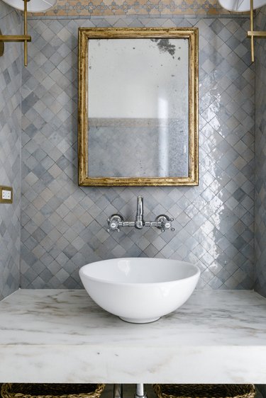 bathroom with blue gray zellige tiled backsplash with traditional mosaic border, brass mirror and sconces and marble vanity