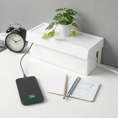 IKEA's Sätting white cable management box rests on a white desk, with a small plant on top of it. It's next to a black alarm clock, with a small notebook in front of it. There is a black cord coming out from the top, connected to a black cell phone.
