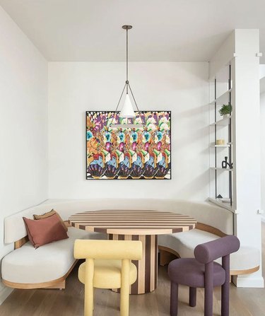 Modern banquette dining nook with striped round table, off-white seating, modern dining chairs, artwork, pendant light.