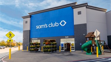 The exterior of a Sam's Club. There are two shelves stocked with flowers right outside the front door and a children's playset off to the side.