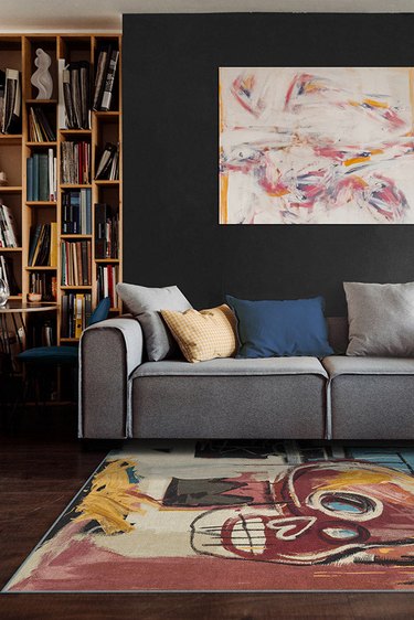 A living room with a grey couch, an abstract painting on a black wall, and a Jean-Michel Basquiat Samo Skull Multicolor Rug from Ruggable on the floor.