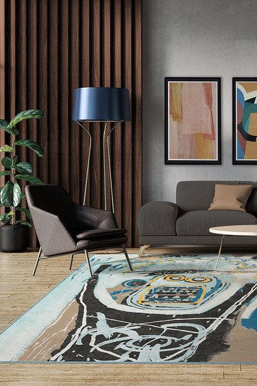 A living room with a dark gray couch and chair, a standing lamp with a dark blue shade, abstract art on the wall and a Jean-Michel Basquiat Ruggable rug on the floor.