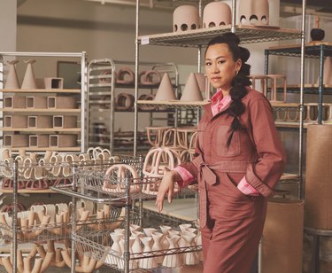 Virginia Sin, a Chinese American woman, wearing a pink jumpsuit and standing in her ceramics studio