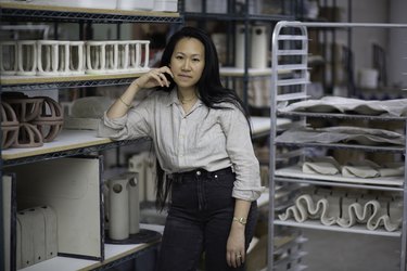Virginia Sin, a Chinese American woman, wearing a gray top and black jeans, standing in her ceramics studio