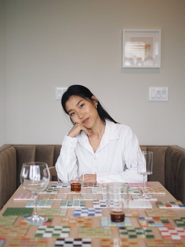 Grace Kim, an Asian American woman, wearing a white button-down shirt sitting at a table covered in her handmade tiled coasters