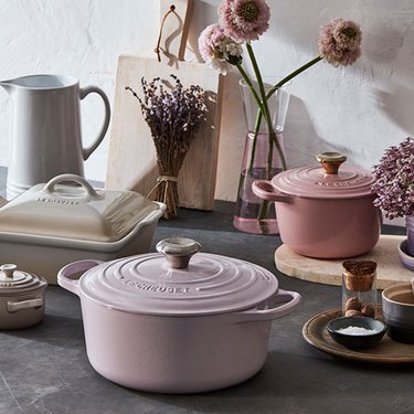 A Le Creuset lavender dutch oven rests on a kitchen counter amidst flowers, a mini dutch oven, a pitcher and a cutting board.