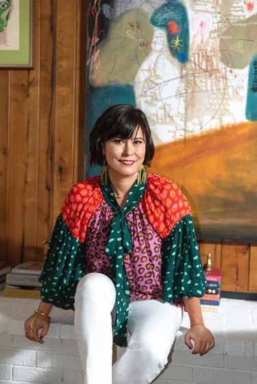 Jessica Davis, an Asian American designer, in a printed shirt and white pants sitting in front of a painting