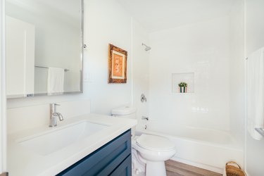 Minimalist bathroom with white walls, counters, blue vanity cabinet, framed portrait, and shower niche with a plant