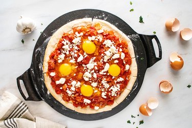 Pizza crust on a cast iron dish with tomato sauce, eggs, and feta cheese