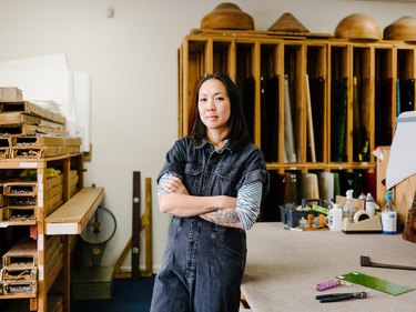 Asian woman with medium-length black hair with tattooed arms wearing black top with blue and white striped sleeves in art studio with cases of glass next to and behind her