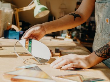 Hands at light wood desk with stained glass pieces with sun shining through circular piece in clear, white, orange, and blue shapes. The person's arms have tattoos and the right hand is holding up the piece.