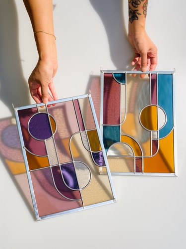 Two hands hold a letter-size stained glass piece in each hand with different-size shapes overlapping in various colors with reflecting shadows in white room