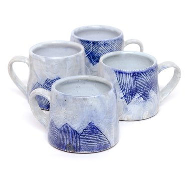Set of four grayish-white mugs with strong blue sgraffito lines with mountain motif over white slip wash against white backdrop