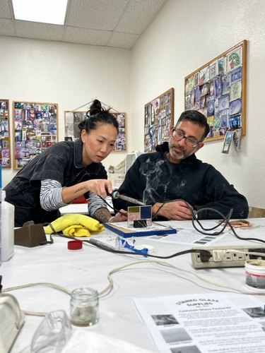 Asian woman with black hair pulled back is wearing a black top with blue and white striped sleeves underneath. She is demonstrating how to cut glass to a man with medium brown skin, short black hair, glasses, and a gray/black beard. He is wearing a black jacket and watching intently. Bulletin boards hang on the wall with colorful photographs.