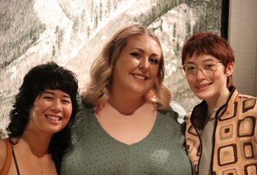 Three women smiling in front of framed illustration in tan and gray: Asian woman with curly black hair and bangs wearing tank top next to medium-length blonde Caucasian woman wearing green shirt and short red-haired Caucasian woman wearing glasses and patterned jacket