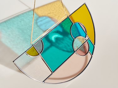 Half-circle stained glass piece with circles and rectangles overlapping in various colors with hanging cord and a reflecting shadow