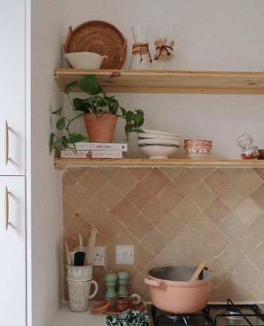 A pink Perfect Pot rests on a stovetop with the lid off and a wooden spoon inside. There is a light pink backsplash and open wood shelving with dishes, a plant, a basket, and two Chemex pour-over coffee makers.