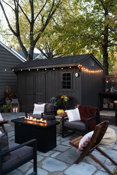 outdoor space with fire pit and chairs near shed in dark color with string lights and trees in the background