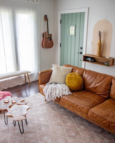 A brown leather couch styled with throw pillows and a blanket on a patterned light pink rug. A shelf and guitar hangs on the wall, and the door is painting a light green.