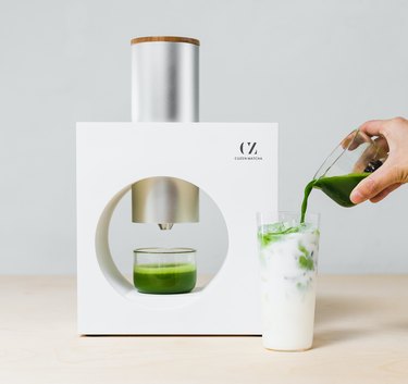 white matcha machine next to glass with milk and person pouring matcha inside
