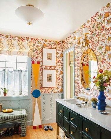 Bathroom with pale blue geometric wainscotting, red and yellow floral wallpaper, forest green vanity, and striped green and white bench