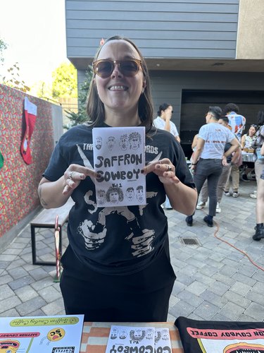 person holding a zine that reads "Saffron Cowboy" with a group of people in the background