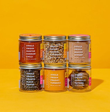 A collection of six Diaspora Co. spices stacked together in two rows. Their labels read Pragati Turmeric Aranya Black Pepper, Guntur Sannam Chillies, Anamalai Nutmeg, Nandini Coriander, and Makhir Ginger.