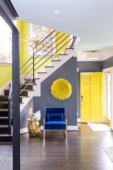 Foyer painted in charcoal and yellow, with royal blue chair, yellow wall hanging, and gold accents