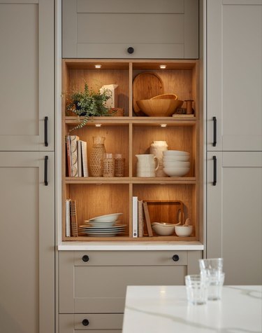 An integrated cupboard with two shelves and in-cabinet lighting. The cabinet is full of bowls, plates, pitchers, books, and a plant.