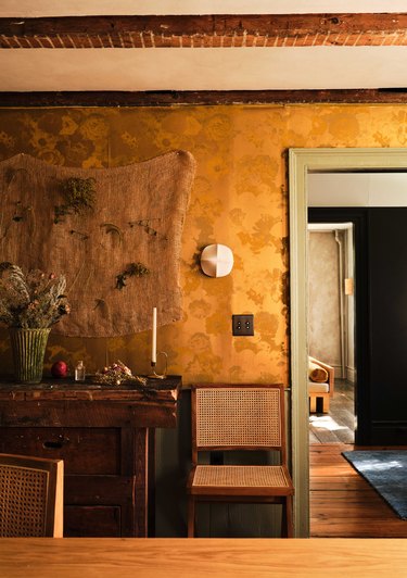 A piece of burlap with dried flowers hangs as art over the credenza with gold wallpaper as its backdrop