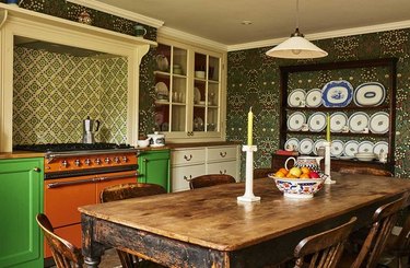 Maximalist kitchen with orange range, green cabinets, rustic dining table, dish rack, glass cabinet