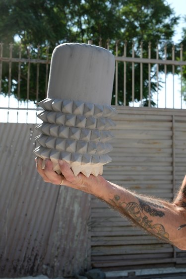 person holding ceramic piece in an outdoor setting with fence and tree in the background