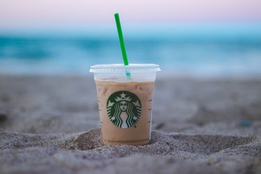 A Starbucks iced coffee drink stuck in the sand with the ocean in the background.