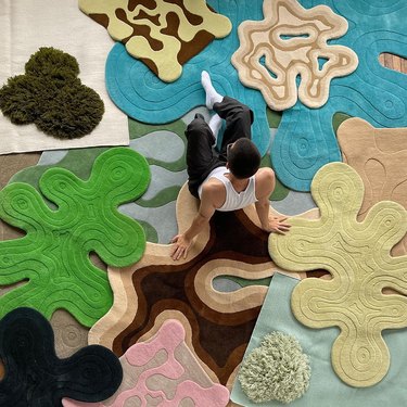 A person sitting on the floor surrounded by irregular-shaped rugs in different sizes, shapes, and colors.