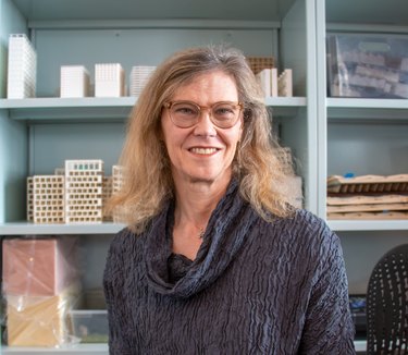 Architect Helen Bronston with shoulder-length blonde hair wearing a pair of light brown glasses and a dark blue, cowl-neck shirt in front of a light blue bookcase.