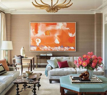 Living room with linen walls, orange artwork, brass chandelier, ottoman, couches.