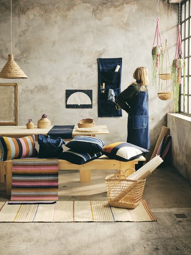 A blonde person standing amidst handmade home decor: throw pillows, rugs, baskets.