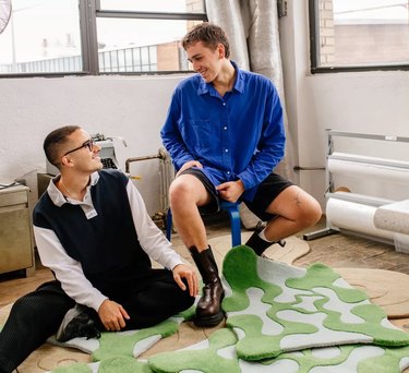 Two men sitting in a room smiling at each other with colorful rugs beneath them