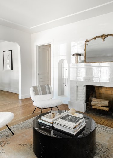 Living room with white fireplace, vintage mirror, white accent chairs, black round coffee table, rug, wood floors.