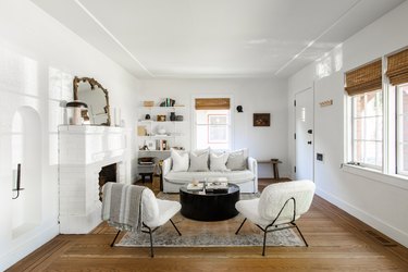 Living room with white walls, fireplace, wood floors, white accent chairs, couch, round black coffee table.