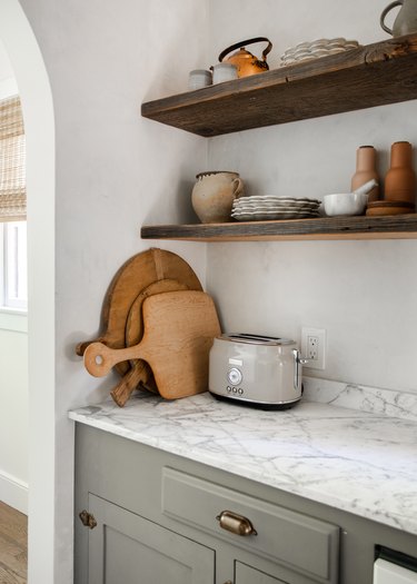Open kitchen shelves with pottery, dishes, teapot, chopping boards, toaster, marble countertop.