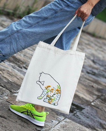 legs and person's hand holding a tote bag with embroidered cat pattern