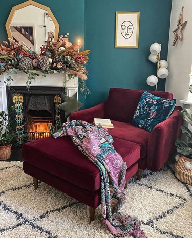 The corner of a living room with teal walls, a plush plum chair, and fireplace.