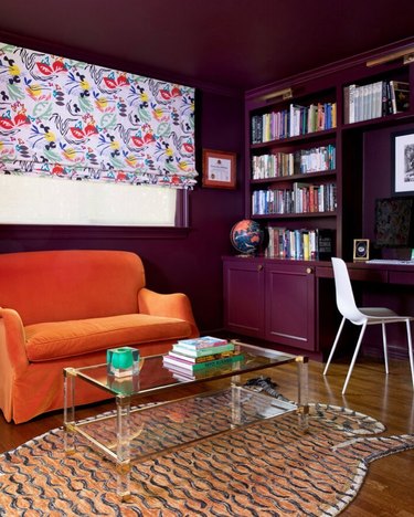 A study with an orange velvet chair, dark plum walls, and a desk area with books.
