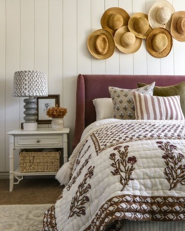A bedroom with a plum headboard, nightstand, and straw hats hanging on the wall.