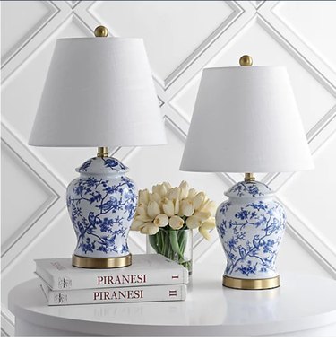 two chinoiserie lamps with white shades on a white table accessorized with books and yellow tulips